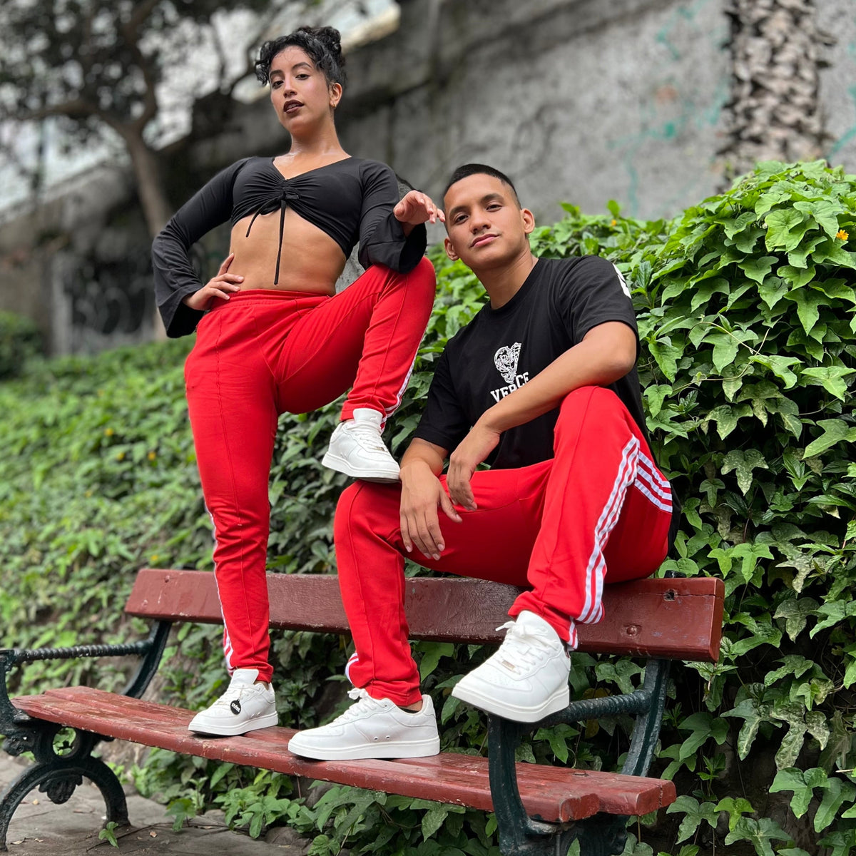 Two dancers on a red park bench by an ivy wall, both in red track pants and white Pulse Dance Shoes dance sneakers